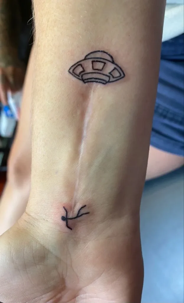 Tatouage sur cicatrice représentant un ovni aspirant un humain, sur l'avant-bras